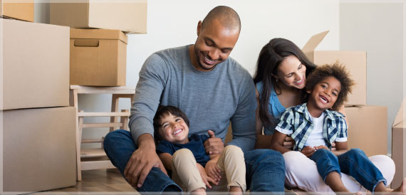 A family of four joyfully sit in their new home with boxes surrounding them. 