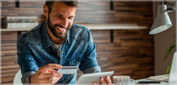 Happy young man sits at his desk with tablet and credit card in hand.